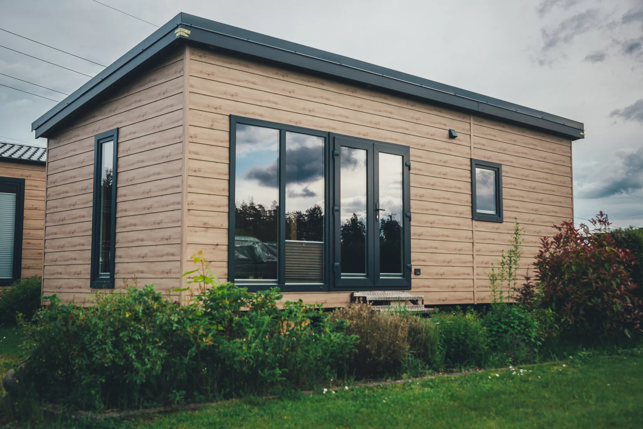 Tinyhaus mit dunklen Fensterrahmen und Terrasse, aufgenommen bei bewölktem Himmel in einem Garten.